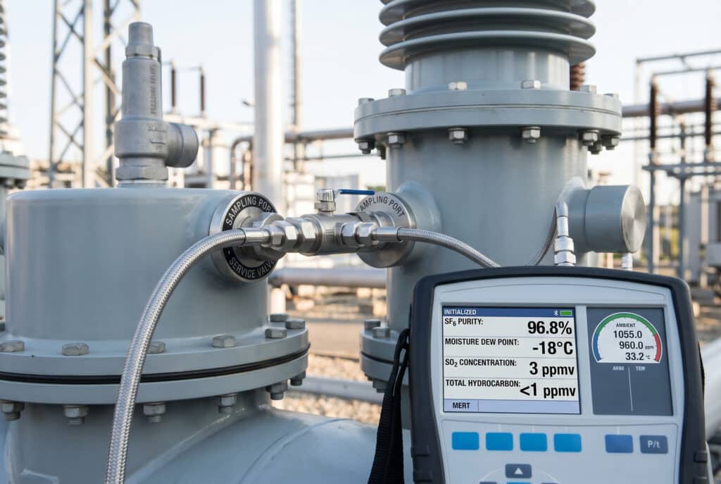 Detailed photograph of a multi-parameter SF6 gas analyzer connected via flexible tubing to the sampling port (service valve) of a large, grey SF6-insulated switchgear or transformer bushing in an industrial plant. The analyzer displays initialization data for SF6 purity, moisture dew point, SO2, and total hydrocarbons, showing real-time measurements from the connected equipment. The focus is sharp on the connections and the digital readouts. Background plant structures are blurred. No humans.