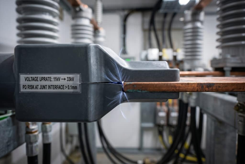 Macrophotographie de l'interface d'un joint de barre omnibus dans une salle de distribution haute tension lors d'une mise à niveau du réseau, où une faible activité de décharge partielle est visualisée à travers les interstices microscopiques et les géométries de détente d'un support d'isolation moulé et d'une barre omnibus en cuivre existante, impliquant une section nouvellement sous tension après l'augmentation de la tension. Une plaque indique "Augmentation de tension : 11kV -> 33kV" et "Risque de DP à l'interface JUNT > 0.1mm".