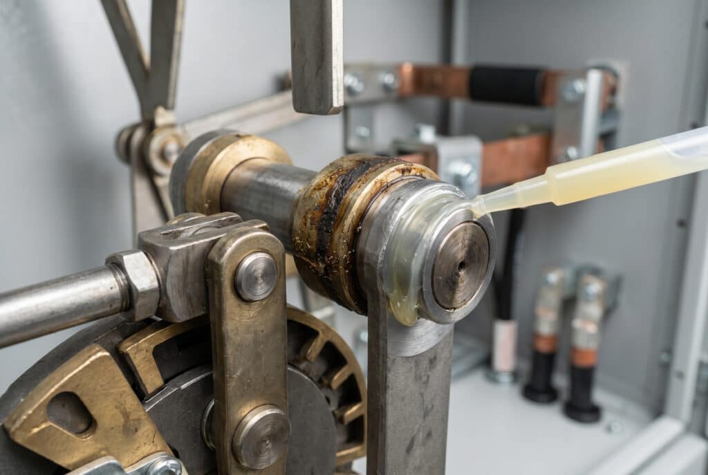 Bir iç mekan ayırıcı anahtarının karmaşık mekanik bağlantıları içindeki aşınmış bir pivot noktasına ve yatak tertibatına uygulanan özel yağlayıcıyı gösteren odaklanmış, teknik bir görüntü, güvenilir elektrik yalıtımı için hassas bakım gerektiren yerel aşınma ve kirlenmeyi vurgulamaktadır.