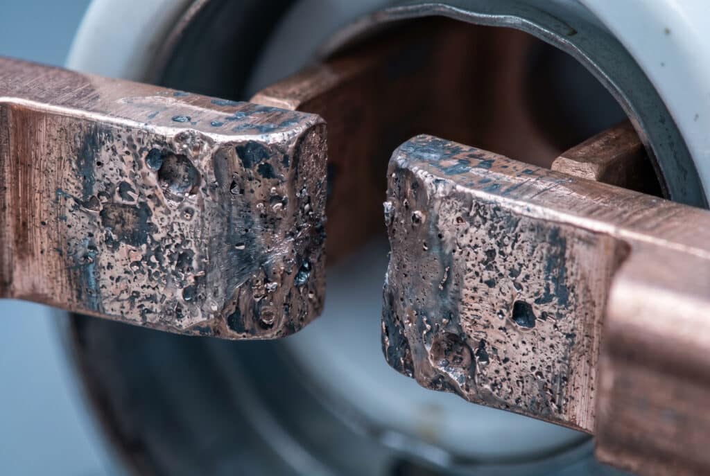 真空遮断器内部の浸食された銅-クロム接触面の詳細なクローズアップ。電気アークによる著しい材料劣化、孔食、摩耗パターンを示し、接触浸食の概念を示している。.