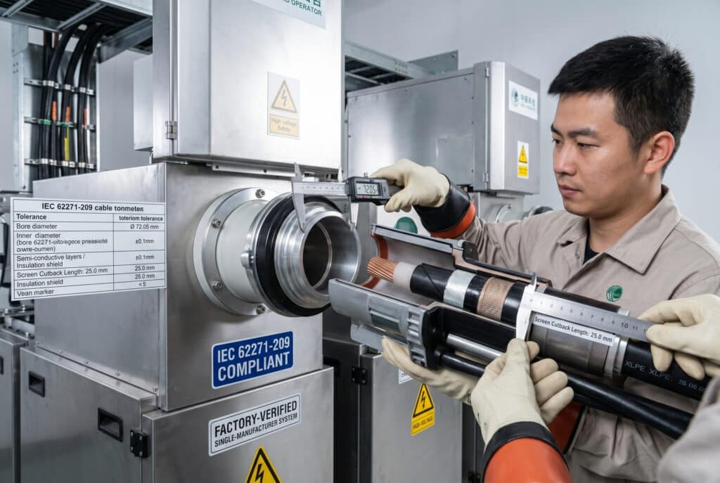 Detailed engineering-style photograph capturing the measurement of a GIS cable termination interface system in a professional substation. A precision digital caliper verifies the bore diameter of a 35kV XLPE cable plug-in connector against the IEC 62271-209 specifications, highlighting 'Bore Ø 72.05 mm' and tolerance compliance (±0.1 mm). Prominent integrated labels read 'IEC 62271-209 COMPLIANT' and 'FACTORY-VERIFIED SINGLE-MANUFACTURER SYSTEM'. Another technician's gloved hand measures screen cutback length on a prepared XLPE cable. The background shows complex GIS switchgear enclosures and cable infrastructure.