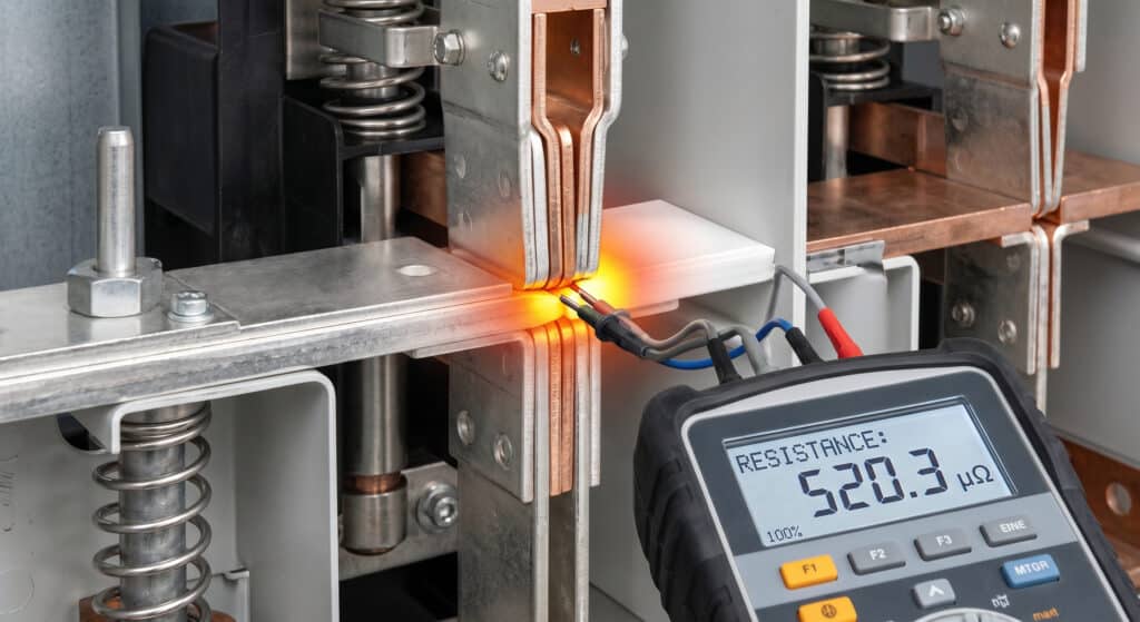 Fotografía enfocada que ilustra el concepto de resistencia de contacto en aparamenta de media tensión AIS, mostrando un conjunto de contacto de cobre cerrado sometido a simulación de calor extremo mientras un microohmímetro mide un valor de resistencia elevado.